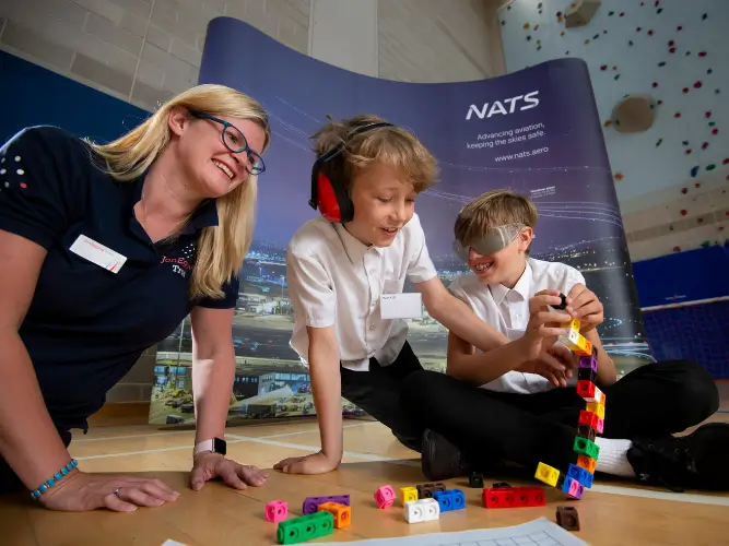 Two students (one with nose cancelling headphones, one with opaque glasses) at NATS doing an activity with JET staff
